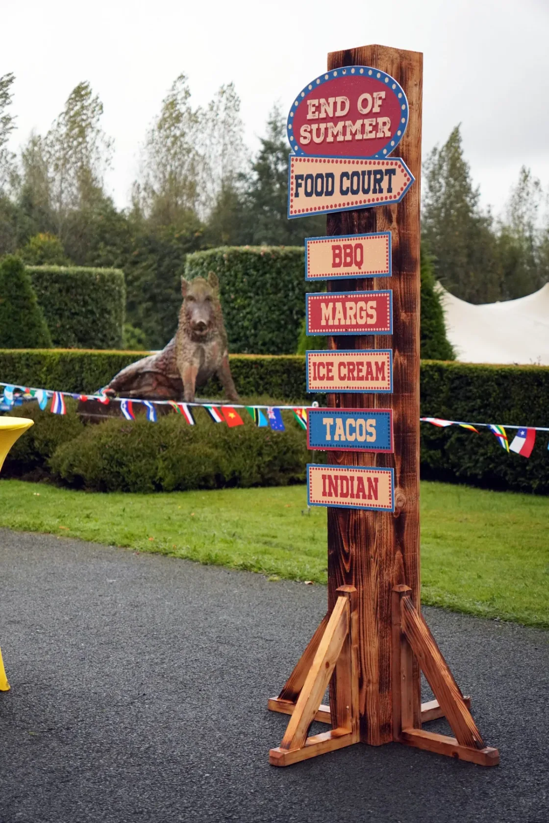A tall wooden directional sign beside an outdoor event entrance.