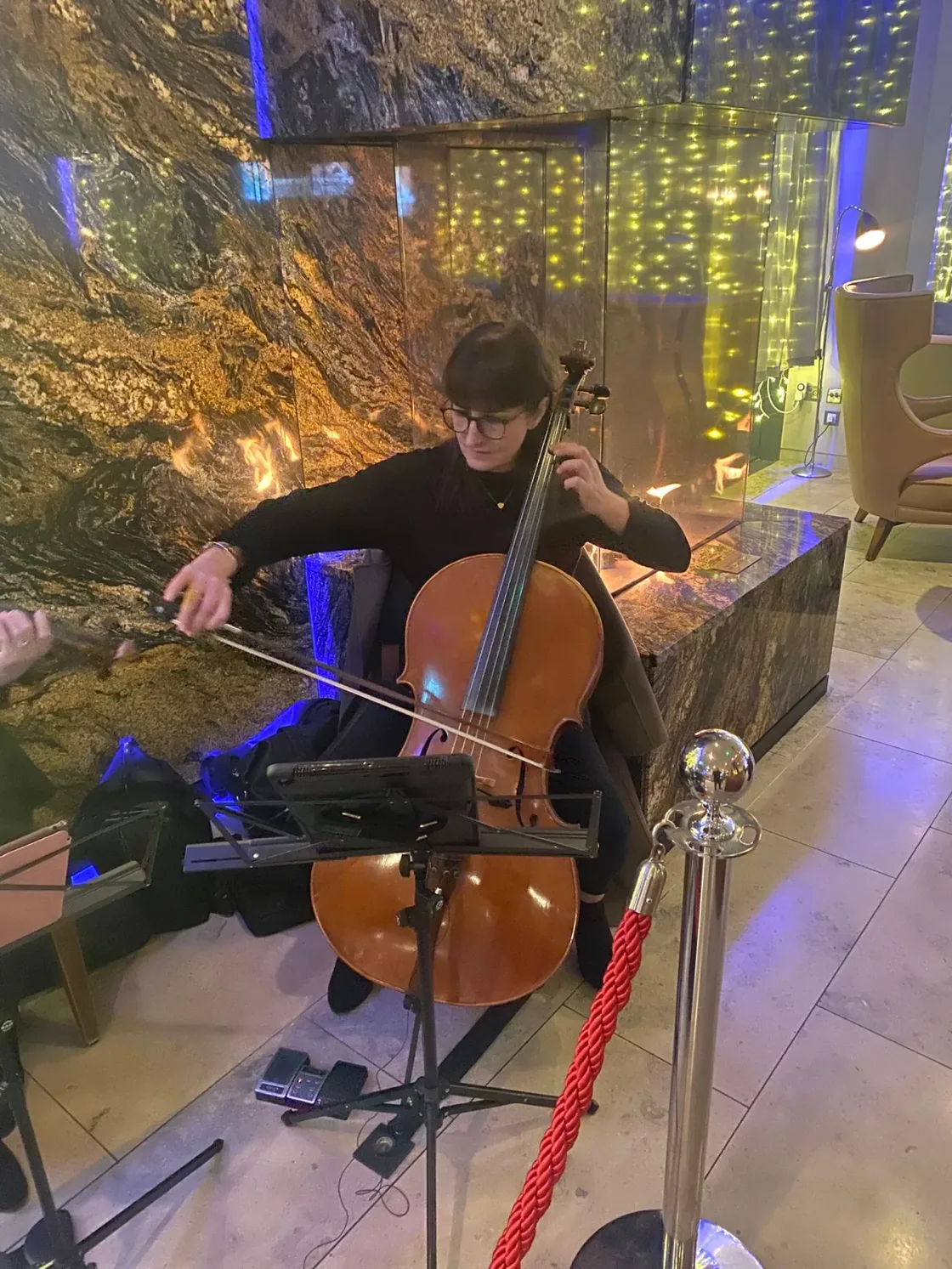 A cellist performing beside foliage and a curtain of warm string lights.