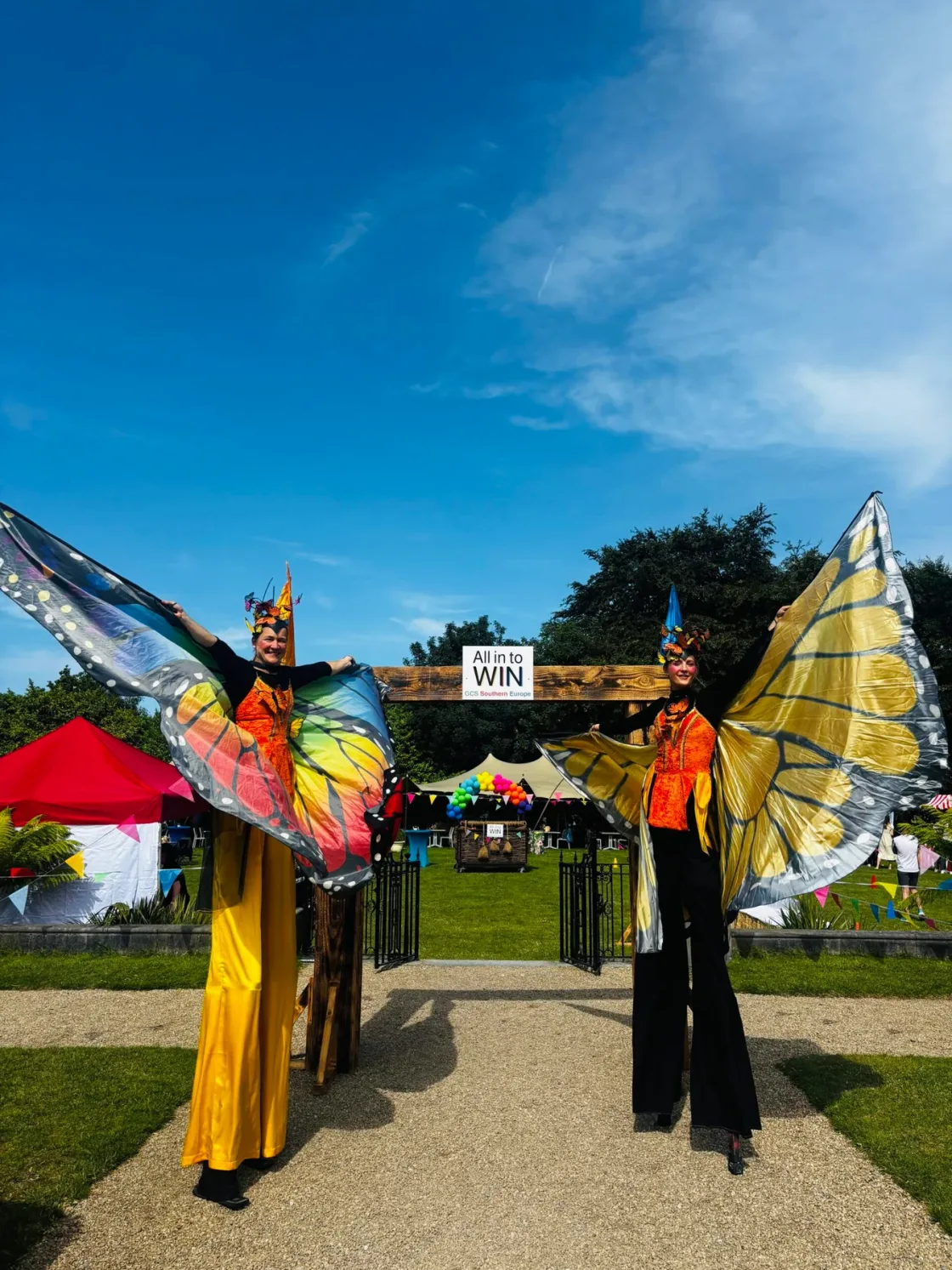 Stilt performers with colourful butterfly wings at an outdoor event entrance.