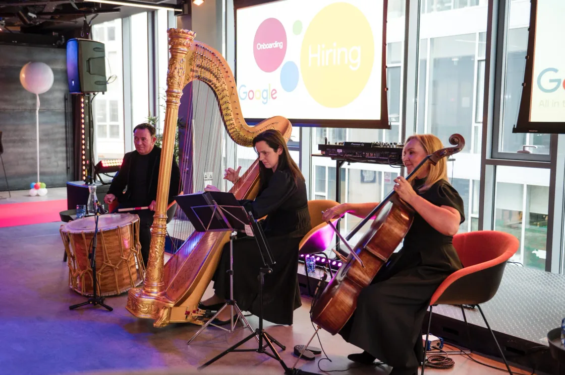 A harpist, cellist and percussionist performing beside presentation screens.