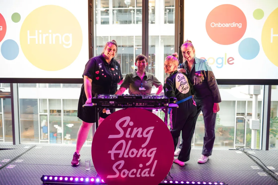 Performers behind a Sing Along Social stand beside presentation screens.
