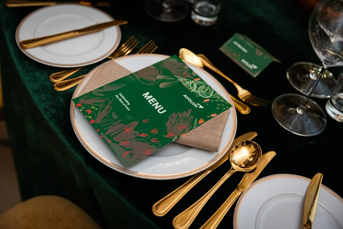 A green printed menu, white plates and gold-coloured cutlery at a dinner place setting.