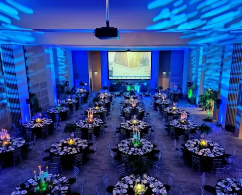 A wide view of round dinner tables facing a stage beneath blue ceiling projections.