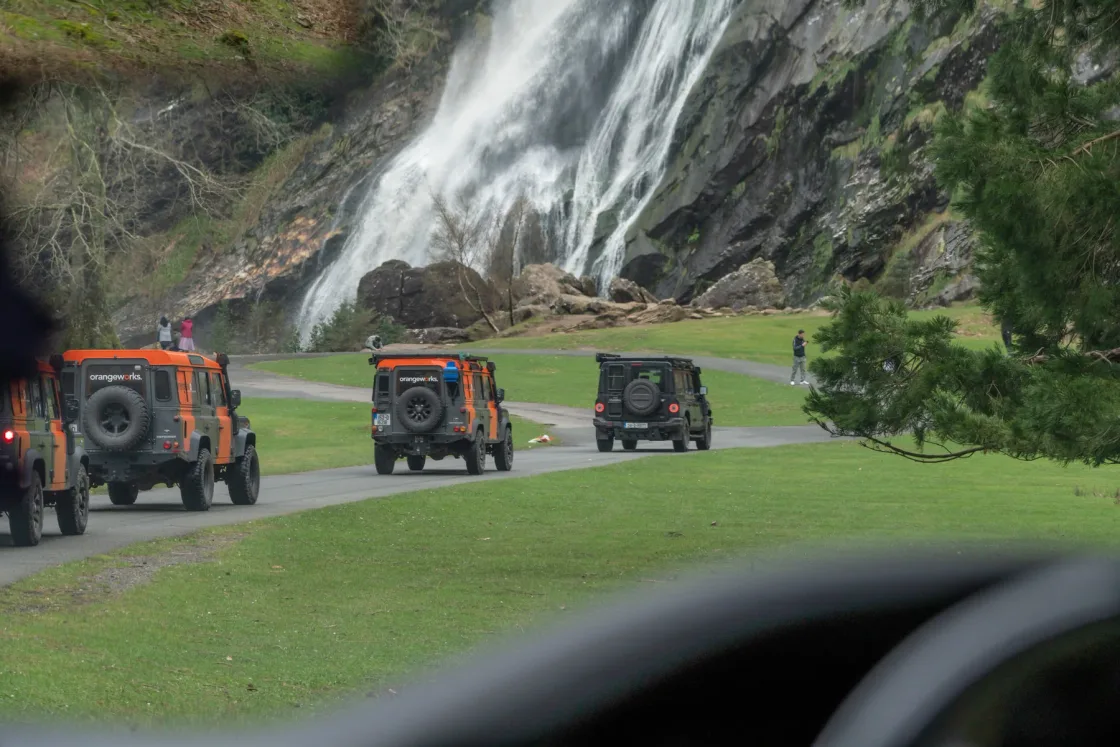Land Rover vehicles parked on grass beside a waterfall.