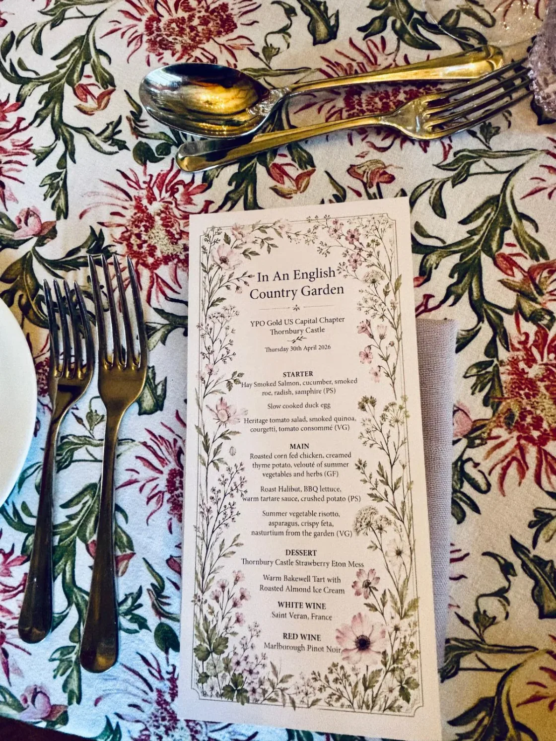 A printed menu on a floral-patterned tablecloth beside gold-coloured cutlery.