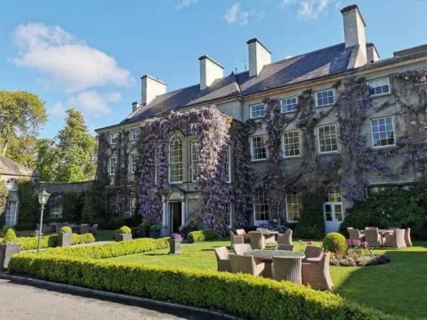 A stone country house surrounded by lawns, hedges and climbing flowers.