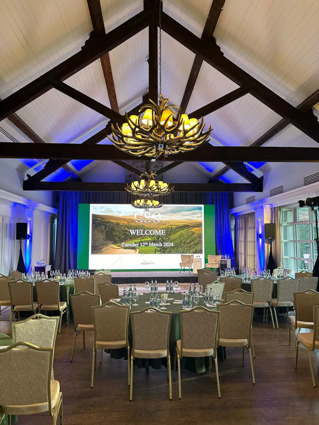 Conference room beneath a timber-beamed ceiling, with round tables and a presentation screen.