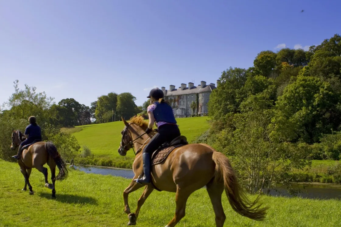 Horse riders crossing a lawn with a large country house in the background.