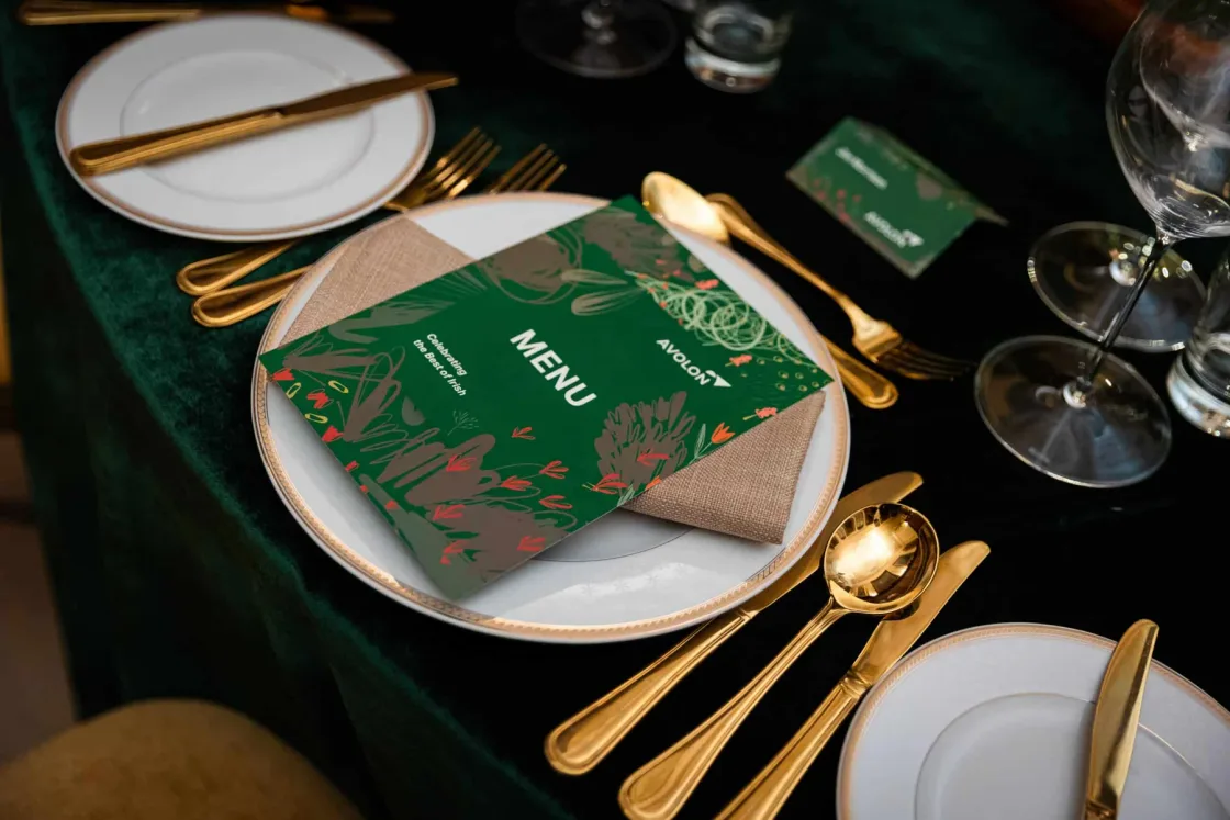 A green printed menu, white plates and gold-coloured cutlery at a dinner place setting.