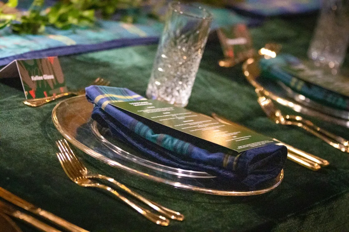 A dark green napkin and printed menu on a glass plate beside gold-coloured cutlery.