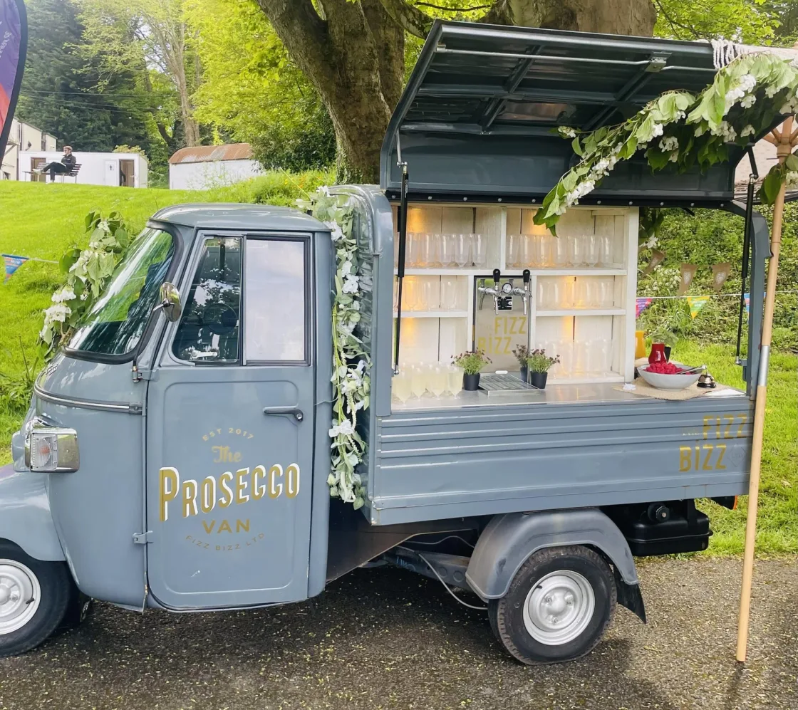 A compact Prosecco van with an open serving counter beside a tree.
