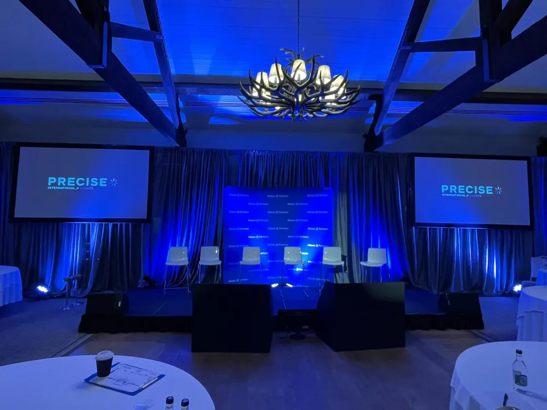 A conference stage lit in blue with PRECISE screens on either side.