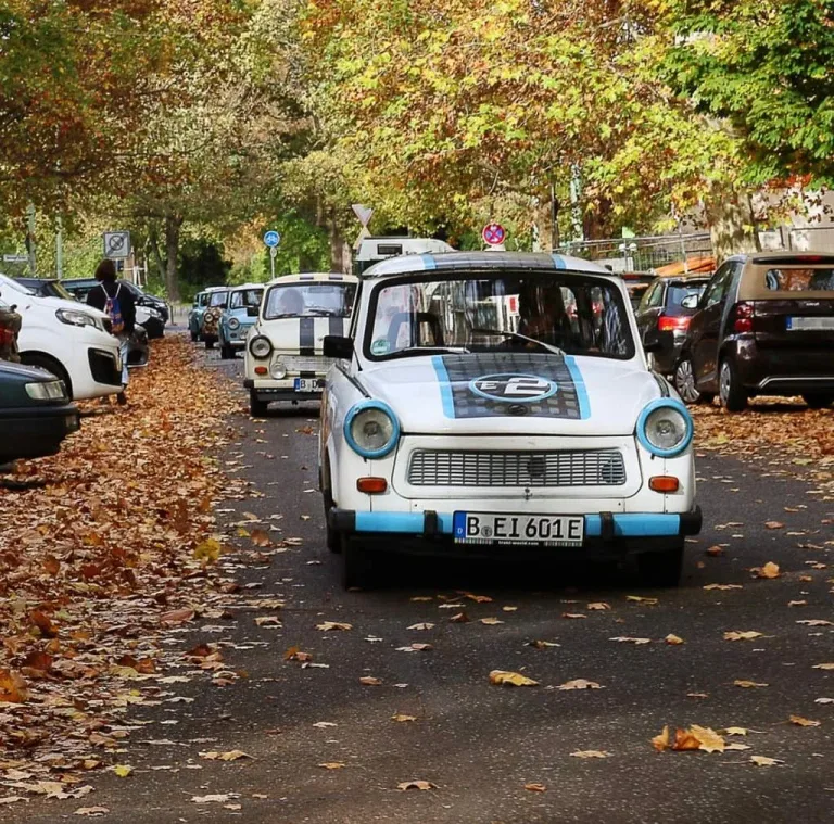 A line of small vintage cars parked beneath autumn trees.