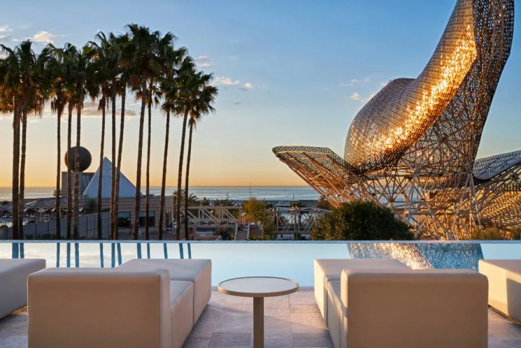 Outdoor lounge seating beside palm trees and a curved metal sculpture at dusk.