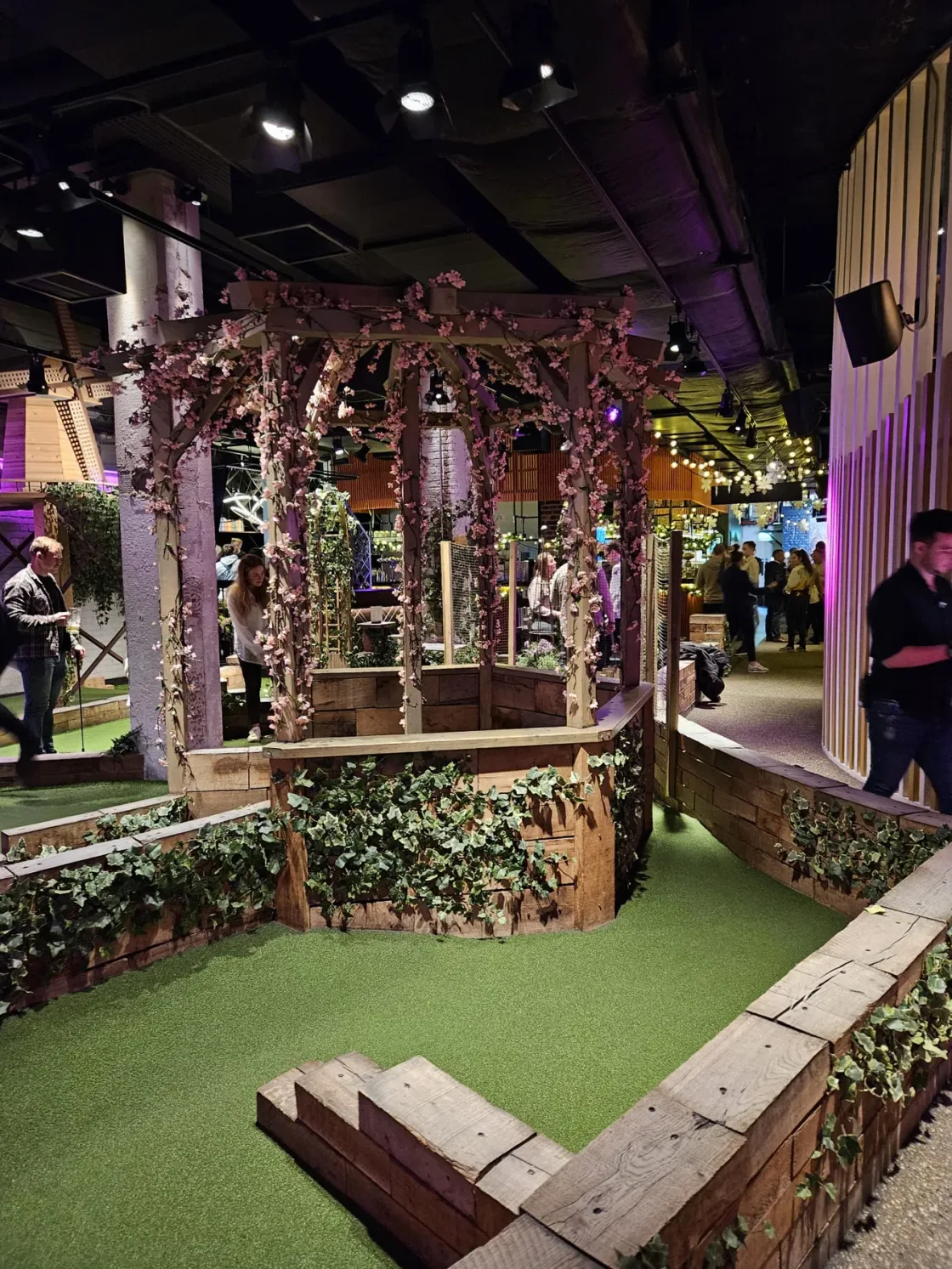 An indoor mini-golf hole beside a decorative hut and planted borders.