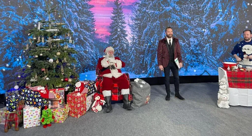Santa and event hosts in front of a snowy backdrop with Christmas trees and presents.