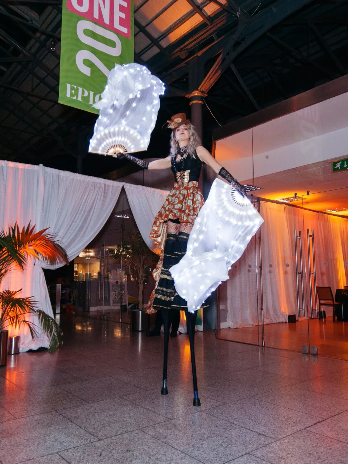 A stilt performer with large white fans inside an event venue.